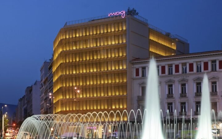 Moxy Athens City’s illuminated facade behind the fountains of Omonia Square at dusk.