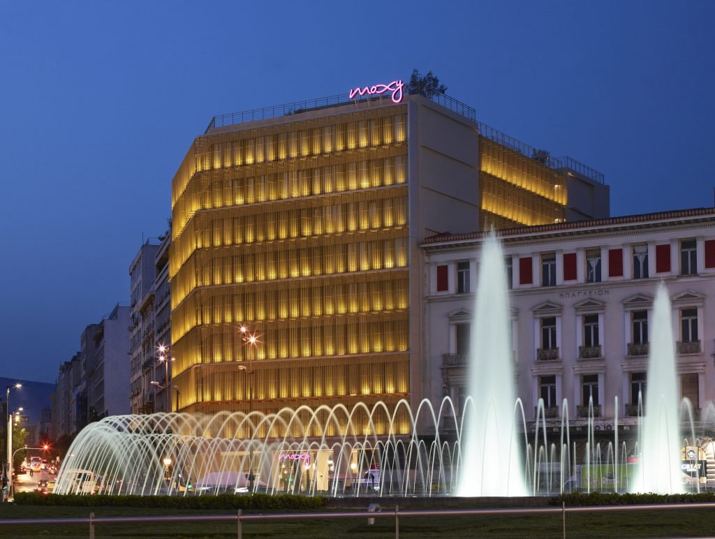 Moxy Athens City’s illuminated facade behind the fountains of Omonia Square at dusk.